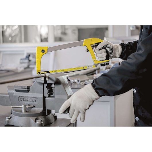 STANLEY 15-098 yellow hacksaw held by gloved hands, cutting metal clamped in a vice, workshop background.