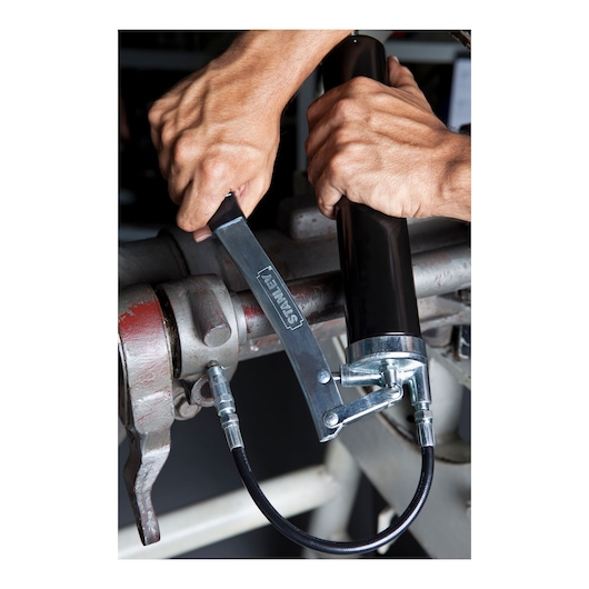 Photo of STANLEY 78-031 grease gun being used on a metal pipe, showing the hand lever and flexible hose.