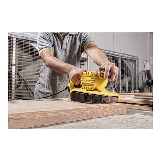 Stanley SB90-B2C belt sander in use on wood, yellow handheld tool, workshop setting.
