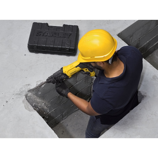 Stanley SHR263K-BR rotary hammer drill in use by worker with hard hat, black case nearby, construction setting.
