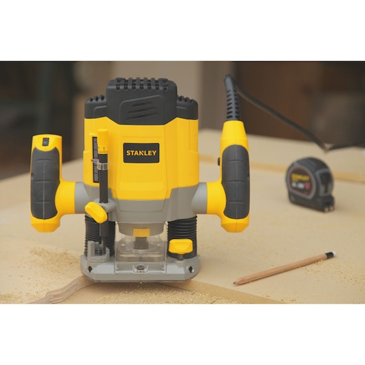 Stanley SRR1200-AR router tool in yellow and black, front angle, placed on a workbench with pencil and tape measure.