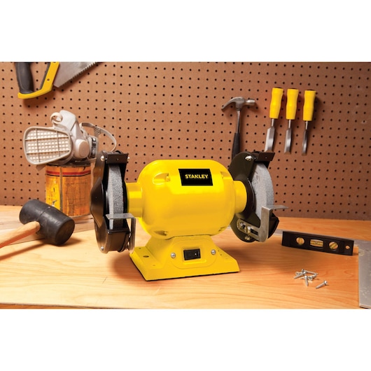 Stanley STGB3715 bench grinder, yellow body, shown on workbench in workshop setting.