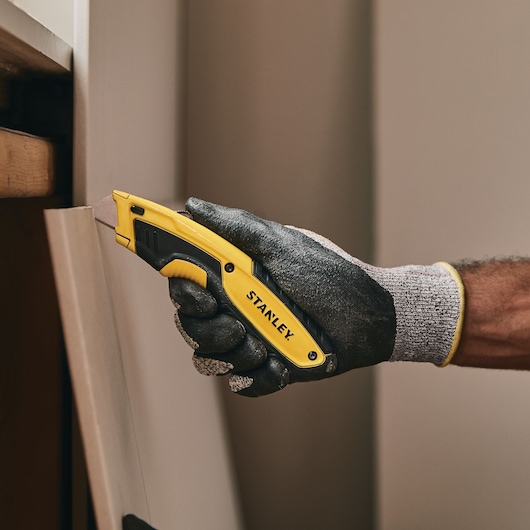 STANLEY STHT10479 utility knife with yellow and black handle, gloved hand cutting wood panel.