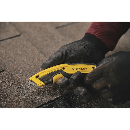 STANLEY STHT10479 utility knife with yellow and black handle in gloved hand cutting roofing material.