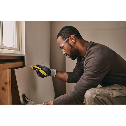 STANLEY STHT10479 utility knife held by a gloved hand cutting drywall in an indoor workspace.