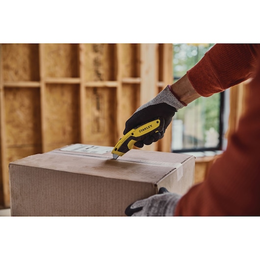 Stanley STHT10479 utility knife cutting a cardboard box, gloved hand holding the yellow tool.