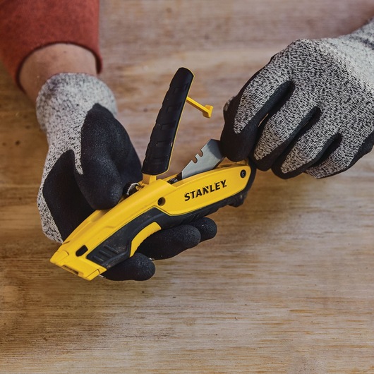 Photo of STANLEY STHT10479 yellow and black utility knife with gloves, shown during blade change on wood surface.