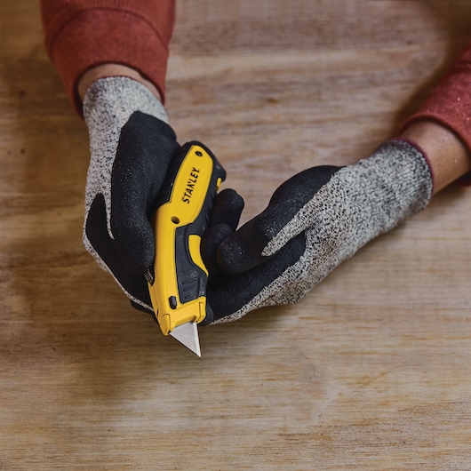 Photo of STANLEY STHT10479 utility knife in gloved hands on a wooden surface, angled front view.