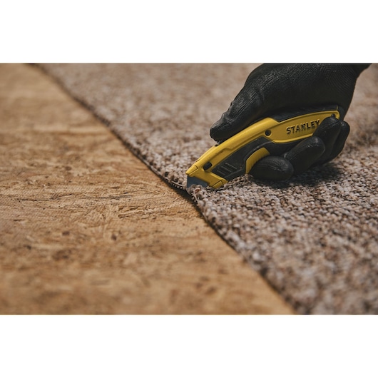Photo of STANLEY STHT10479 utility knife cutting carpet, close-up, black glove holding yellow tool.