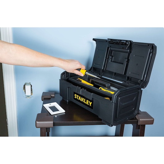 Photo of STANLEY STST16420 black plastic toolbox with open lid, showing hand reaching for yellow tool, on table.