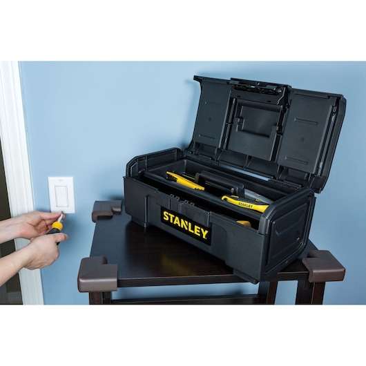 STANLEY STST16420 black toolbox open on table, showing tools inside, person using screwdriver near light switch.