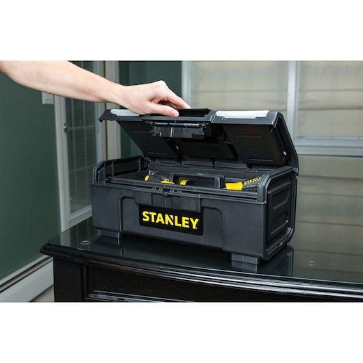 Photo of STANLEY STST16420 black plastic toolbox with top lid open, showing hand lifting lid and tools inside.